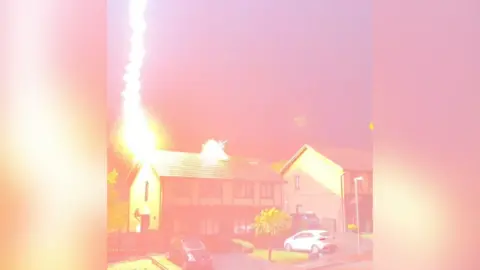 A lightning bolt is seen striking a detached house. The image is tinged with pink and orange capturing the moment the lightning hits the roof of the house. It is in a residential area with trees, cars and another house on the left hand side of the building.