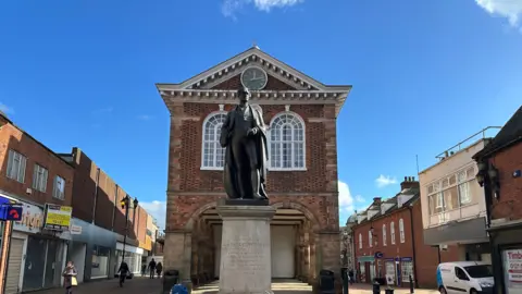 Tamworth Borough Council Tamworth Town Hall, a building with an open space and a series of columns underneath it. There is a statue of Sir Robert Peel in front of the building. 
