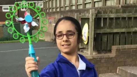 Shay Kang wearing a blue school uniform in what could be her school's playground holding a large, green plastic toy towards the camera. She has dark hair tied back and is wearing glasses.
