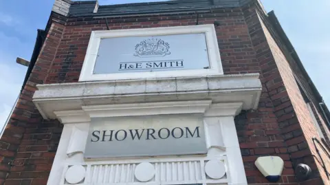 A red brick building, with an off-white stone entrance, bearing a crest, and the words H&E Smith and showroom