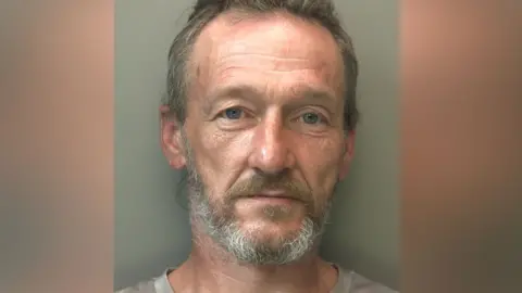 A police mugshot of a man with grey hair and a beard. He is looking directly at the camera.