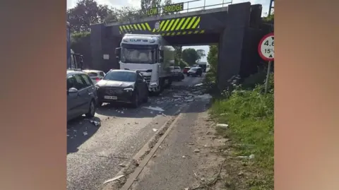 Supplied A lorry underneath the bridge, with an amount of debris on the road. There are cars queued behind and in front of the lorry.
