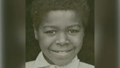 Fumi Stephenson A head-and-shoulders picture of Dr Stephenson as a child. He is smiling at the camera and wears a light-coloured checked collared shirt. 