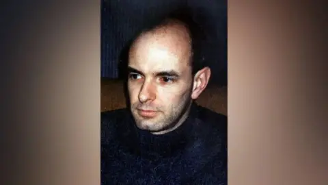 Christopher Farrow looking off-camera, he is balding, with black or dark brown stubble and eyebrows, dark eyes, and tan skin.