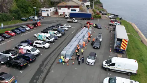 Ben Birchall/PA Wire A shot from above a car park with pallets of water bottles stacked in the middle and cars queueing to collect the bottles.  There are people in high-vis jackets handing out the packs of water bottles. 