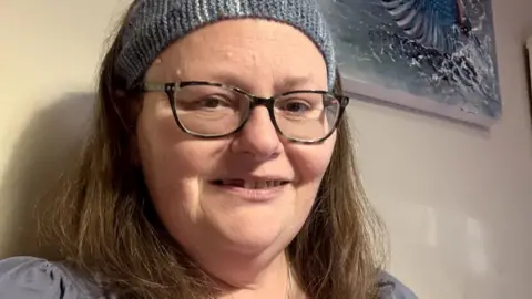 Selfie of Sarah Bodger, who has brown hair and is smiling at the camera. She is wearing a blue knitted hair band, glasses, and a blue top.
