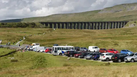 Crowds of people walk to and from the Ribblehead Viaduct, with a busy car park in the foreground. 