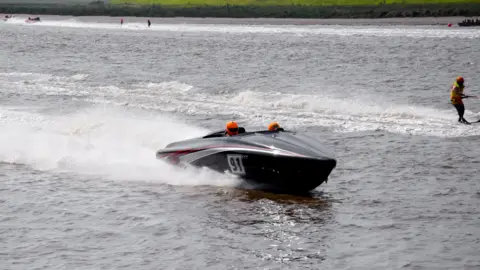 O Conselho de Boro de King's Lynn e West Norfolk viaja para o Great Ous River. O motorista está usando um capacete laranja brilhante. O número 91 aparece na lateral do barco.
