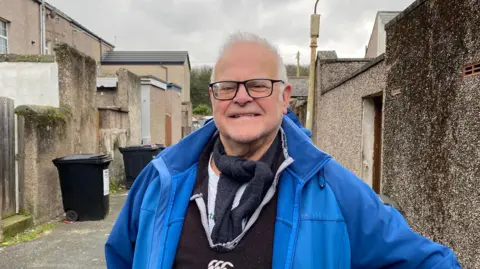 Brian Leeson is standing in a twitchel. He has short white hair and is wearing a black pair of glasses and a blue coat.