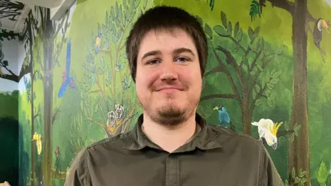 Chris Evans has black hair and trimmed beard. He is wearing an olive coloured shirt and is standing indoors next to a jungle-themed mural.