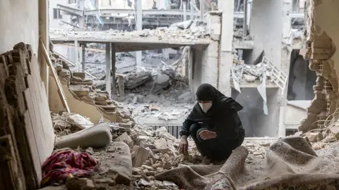 A woman wearing a face mask and dressed in black sifts through the rubble in her house in Tehran after it was damaged by missile attacks. The wall behind her as been completely destroyed, as have other buildings behind it.