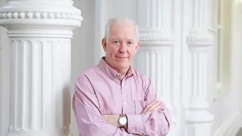 Peter Murphy, de la Universidad Nottingham Trent, está de pie, con los brazos cruzados y una camisa rosa. Está apoyado en una columna blanca. Sonríe y tiene el pelo corto y blanco.