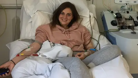 Supplied Holly Murdy, lying but with her back propped up, on a hospital bed. She's wearing a pastel pink hoodie and grey jogging bottoms, has dark shoulder-length hair and is smiling. Her arms have intravenous tubes attached with tape, with yellow, red and green valves. To her left is a piece of hospital equipment, into which some of the tubes are fed.