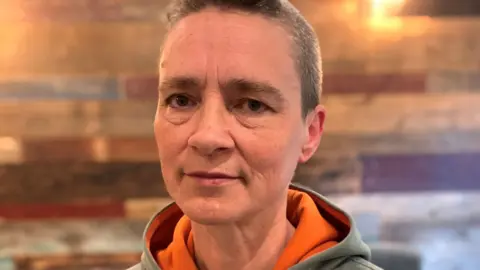 A woman with very short brown hair looks into the camera with a straight face. She wears a grey hoodie with orange inside the hood.