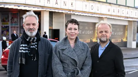 Sam Mendes, Olivia Colman and Toby Jones smiling for the camera at a roadside. Behind them is a cinema building which has "thank you Margate" on a lit sign along the length of the image and two posters for the film Empire of Light on the wall.