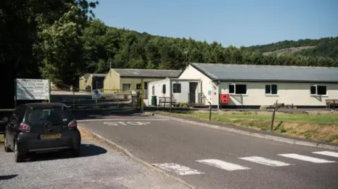 Hammonds Yates A section of the former Yeo Valley Farm site in Cheddar. It shows an entrance driveway with a zebra crossing and a metal gate securing the site. There are long one-storey buildings with corrugated metal roofs. In the distance there are fields and mature trees.