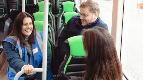 From left to right, Louise Lister, Oxford Bus Company Assistant Marketing Manager and David Moore, Oxford Bus Company Marketing and Events Officer. They are sitting on a bus and speaking to a woman. The woman has her back at the camera. Louise and David are smiling.