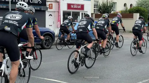 Image of a group of men on bicycles riding on a street lined with cars and shops.