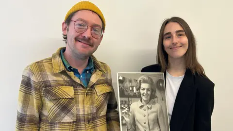Gavin Kermack A young man wearing round, red-rimmed glasses, a yellow beanie hat and a yellow and black lumberjack-style shirt over a blue-green check shirt stands next to a young woman with long brown hair. She is wearing a black jacket over a white t-shirt. They are holding a print of the above picture.