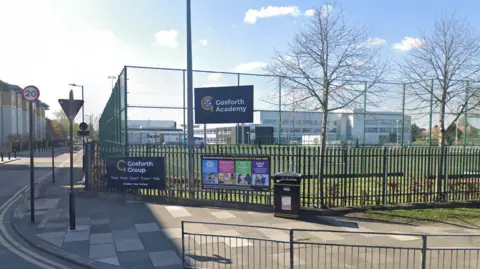 Gosforth Academy. The picture is taken fro the road and shows the school building. A fence has been placed around a sports pitch in front of the school. Two signs displaying the school's name are hung on the fences.