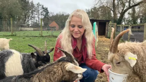 Photograph of Ball, who is wearing blue jeans and a red jumper, crouching down in a field feeding four goats. The goats are eating out of white plastic buckets. The sky is grey and overcast. 