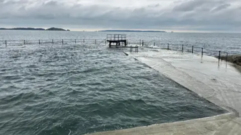 A diving board on the edge of an open air seawater bathing pool on a grey-weather day. Both the sea and pool are at the level of the edge of the pool, and there are islands in the background.