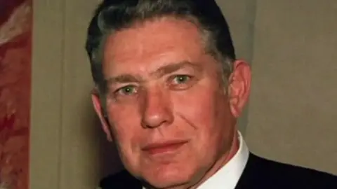 A man with short dark hair looking ahead. He is wearing a white shirt and black jacket. 