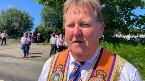 David Mahon, a man with short, fair hair, looks to the camera during a BBC interview on a very sunny day in rural Donegal.  He is wearing a white shirt, a red. white and blue striped tie and an orange sash with a gold fringe. 