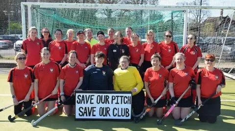 Southwick Hockey Club Southwick's women's team lined up in the goal mouth of their pitch with their hockey sticks and red tops with a sign that reads "save our pitch, save our club".