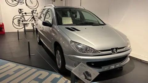 Peugeot 206 built in 2006 at Ryton parked in the Coventry Transport Museum with a display sign about the car and bikes on the wall