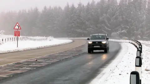 A dark-coloured car drives along a road cleared of snow. A red reduce speed sign is on the opposite side of the road. The sides of the road are covered with snow and black and white bollards.