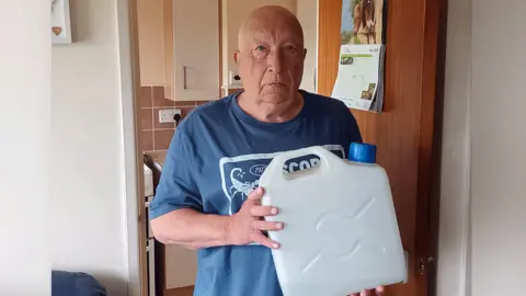 LDRS Richard Tobin with a clear plastic water container used to cope with water pressure problems at Caerau Court in Cardiff. He is bald and wearing a blue t-shirt, standing in a doorway with his kitchen behind him.