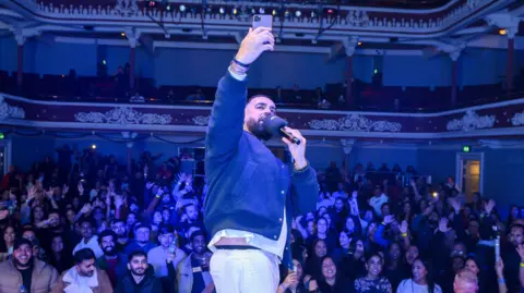 A DJ stood on a stage holding a microphone in one hand and a phone in the other, taking a selfie with the crowd. The audience fills the theatre’s balconies and floor, with many people raising their hands under blue lighting.
