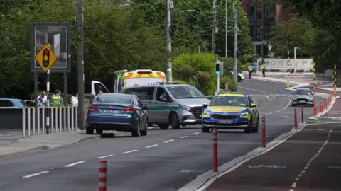The side of a road with a grey ambulance, a blue and yellow garda car, a blue and yellow garda van, a blue can parked on the pavement and another grey car in the distance.