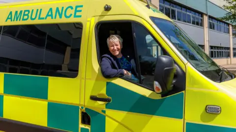 MP Rachel Taylor is sitting at the wheel of an ambulance which is liveried in yellow and green. It is parked outside an office building.