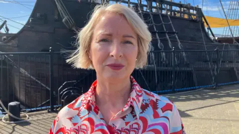 Helena Hasson stands on the riverfront with a replica Spanish galleon in the background. She has blond hair and is wearing a pink and white patterned blouse