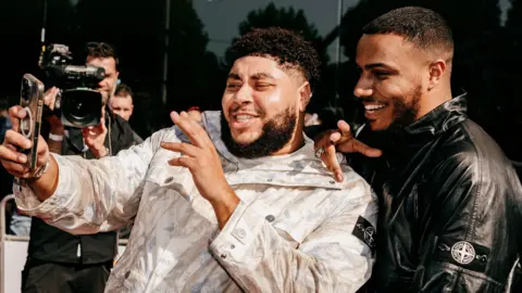 Getty Images AJ Tracey and Big Zuu taking a selfie on a red carpet. AJ (right) smiles over Big Zuu's shoulder as he holds up his phone. AJ wears a black jacket while Zuu is in a white coat. It's sunny outside and members of the media can be seen behind them.