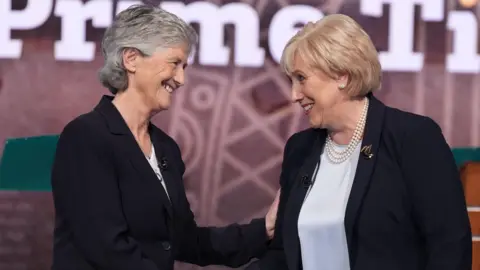 Niall Carson/PA Media Catherine Connolly shakes hands with Heather Humphreys as the two take part in a televised debate. Connolly has short, grey hair and is wearing a dark suit jacket and white shirt. Humphreys has short, blonde hair and is wearing a pearl necklace, a dark suit jacket and a white satin shirt.