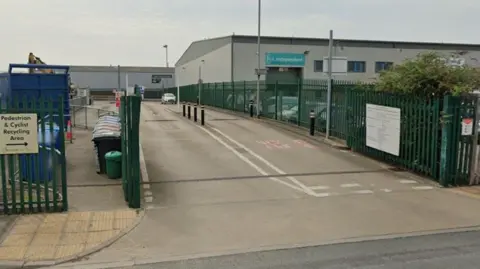 Entrance to Hazel Court tip on James Street in York. There are green railings lining the entrance road, with a sign to the left pointing to a 'Pedestrian and Cyclist Recycling Area'. To the right behind the railings there's a large industrial building, with 'Be Independent' above the doors.