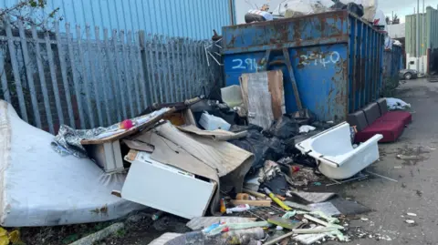 An image of rubbish, including a bath, spread out on a road. A huge, blue metal container is in the background next to a fence.