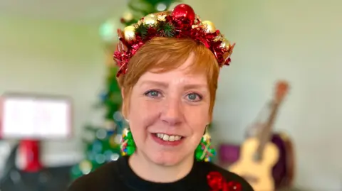 Martin Giles/BBC Helen Barnes is smiling at the camera. She has short ginger hair and is wearing a black jumper, Christmas tree earrings and a headband which is made from tinsel and red and gold baubles.