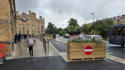 A brick station building with a clock on it. A footpath with people on it and a line of silver bollards. A wooden planter with a no entry sign on it and plants in the top blocks a path next to the bollards. A road next to the path has cars and a bus on it.