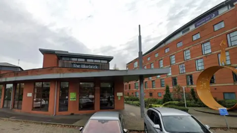 Google The Bluebrick building is on the left, with the sign visible. The building on the right, with five storeys, has a Premier Inn sign near the top. Two cars are parked in the foreground.