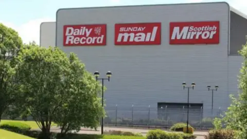 Reach PLC A close up of Reach's Saltire printing facility, which is a big metal-clad industrial building. On its side are big logos for the Daily Record, Sunday Mail and Scottish Mirror. The sun is shining and it is surrounded by trees.