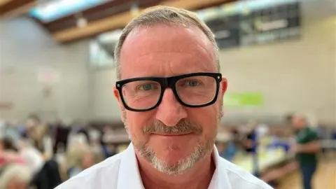 Tim Dwelly wearing thick black framed glasses. He has short brown hair and a stubbly beard. He is stood in a sports hall with counting taking place behind him.