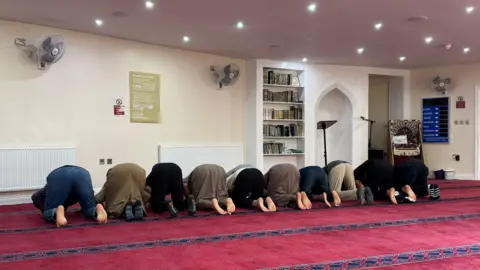 Charlie Rose/BBC On a thick burgundy carpet, praying worshippers are on their knees with their faces to the floor.