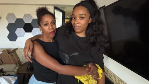 Isabel and Aysha stand indoors with their arms around each other. Isabel, wearing a black top and yellow scarf, looks towards the camera while the Aysha, in a sleeveless black top and light blue trousers, leans into her. Behind them are a wall-mounted television, hexagonal wall panels, a keyboard, and cardboard boxes on a sofa.