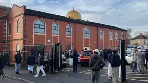 People dressed in traditional dress surrounding a a modern red-bricked mosque building