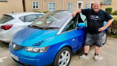A man in a black t-shirt, navy shorts and trainers is stood next to a blue and silver Renault Avantime. He has one arm on resting on the roof and a design of the car is printed on his shirt. The car is parked beside a smaller silver car outside a modern brick building.