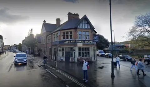 Google The front of the Cape of Good Hope pub on a damp day. There are some people walking in front. A car is driving by on the street. There is a man riding a bicycle by the pub.